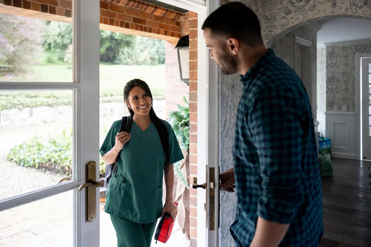 A phlebotomist in green scrubs greeting a patient at the front door of a Portland home